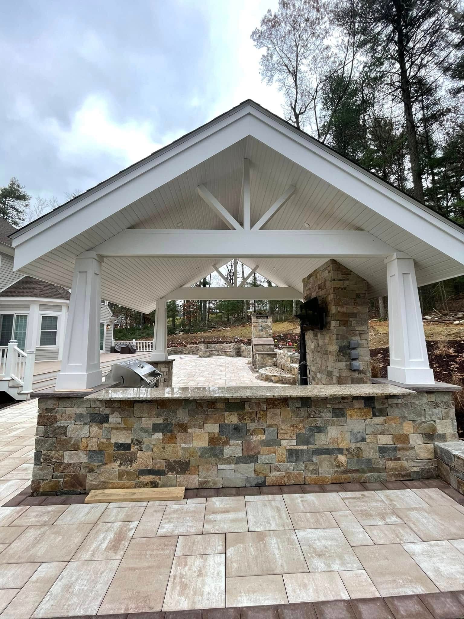 Outdoor kitchen bar clad in natural stone veneer with a granite counter, built-in grill and open gable pavilion above