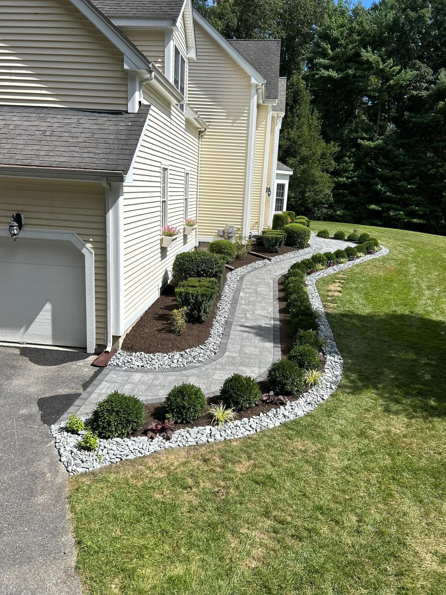 Paver walkway running from the driveway along the house, bordered by clipped boxwood and a white stone drainage margin