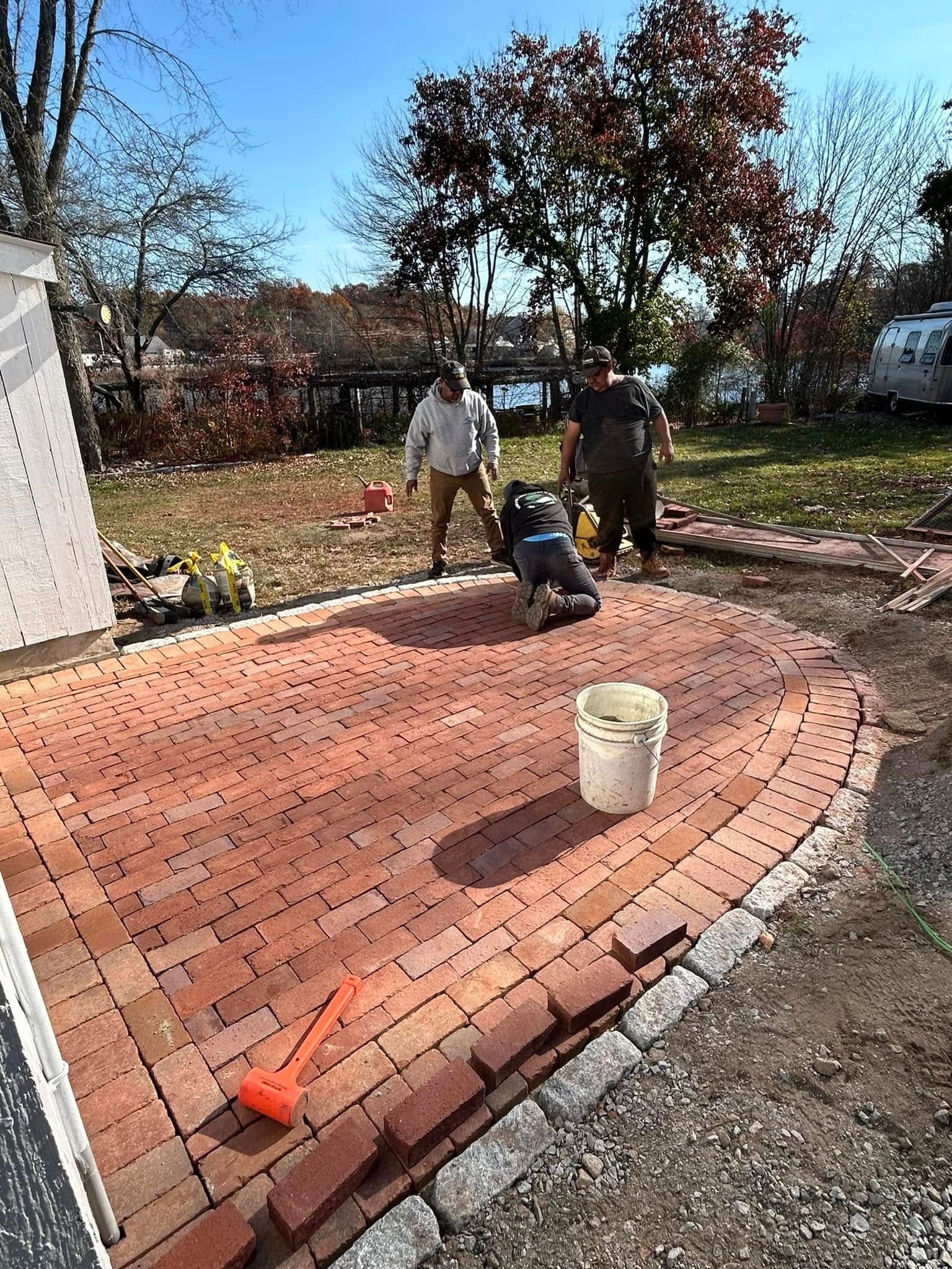 Three-man Kamaye crew laying a radial reclaimed-brick terrace with a granite cobble edge restraint, water beyond