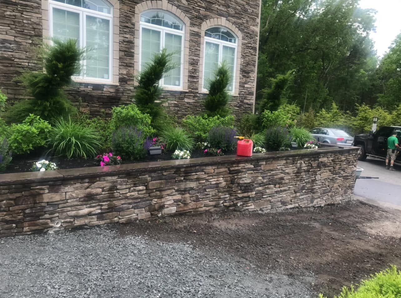 Dry-stack stone retaining wall with a bluestone cap holding a planted bed above a driveway base being prepared
