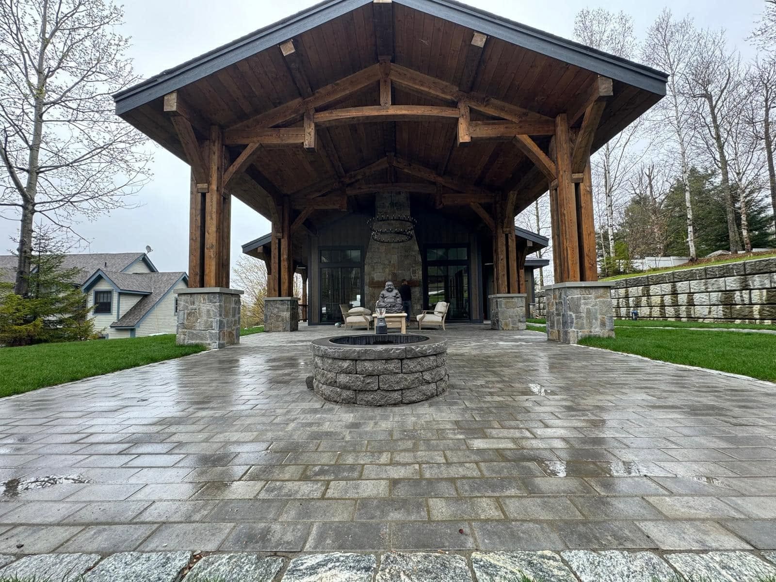 Timber-frame pavilion with a stone fireplace above a wide paver courtyard and a circular granite fire pit, southeastern Massachusetts